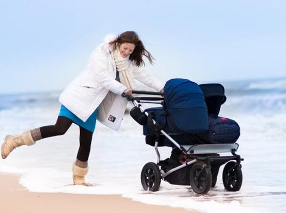 A mom playing in the water on the beach in winter while pushing a double baby stroller.