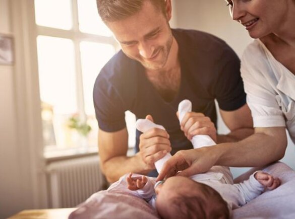 A father plays with his newborn baby while the mother looks on smiling.