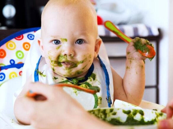 A parent feeds his baby in a highchair with green vegetables as baby food.