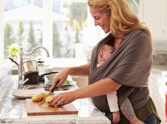 A mother prepares a healthy meal to support good nutrition during breastfeeding while her baby sleeps in a sling attached to her.