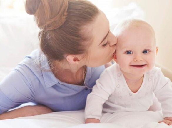 A baby crawls around on the bed while its mother lies next to it and gives it a kiss on the side of the head.