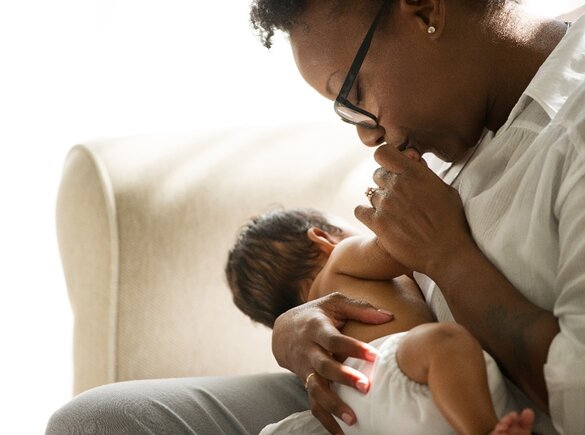 Woman sitting on a couch holding a newborn baby and kissing the baby's hand.