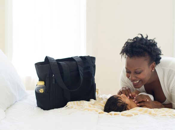 Baby laying on bed next to a Medela pumping bag. Woman is smiling down on baby.