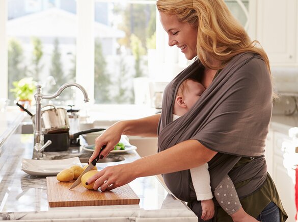 A mother prepares a healthy meal to support good nutrition during breastfeeding while her baby sleeps in a sling attached to her.
