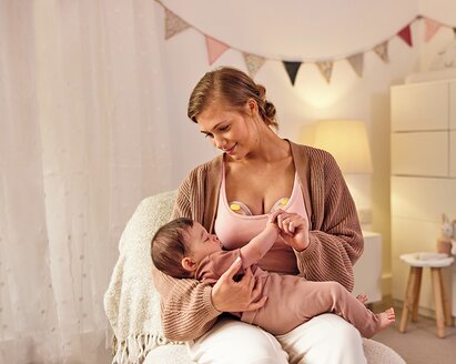 Eine Mutter trägt eine elektrische Medela Swing Maxi™ Doppelmilchpumpe, während sie ihr Baby im Kinderzimmer hält.