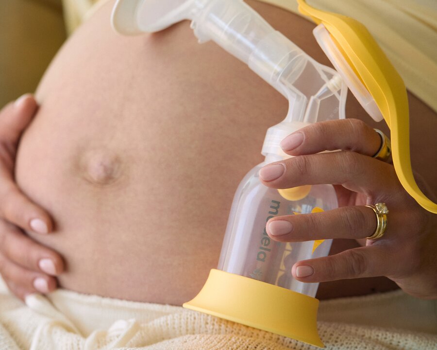 Close-up of a person holding a Medela Harmony manual breast pump against the abdomen. The yellow handle, breast shield, and collection bottle of the manual pump are visible, with the product held in one hand while seated in light-colored clothing.