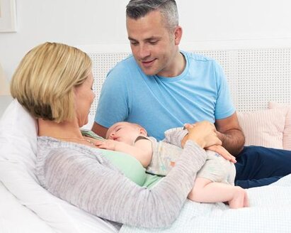 A mom lies on the bed while the baby sleeps on her chest, the dad sits on the bed next to them.