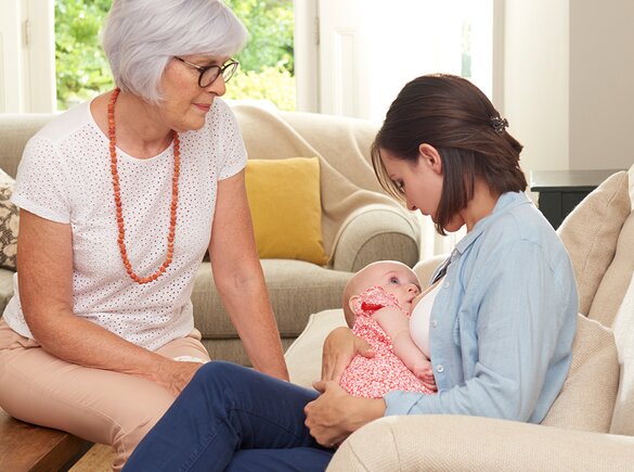 Breastfeeding A mother breastfeeds her baby at home in the living room while the baby's grandmother supports her.