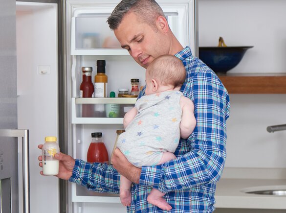 Un padre tiene in braccio il suo bambino in cucina mentre prende il biberon dal frigorifero per riscaldarlo per la poppata.
